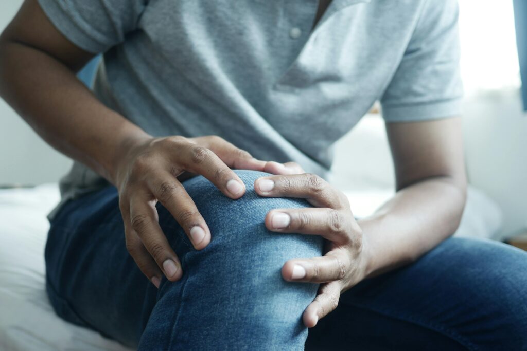 Sitzender Mann greift sich an sein rechtes Knie mit beiden Händen weil er Gelenkschmerzen hat. 